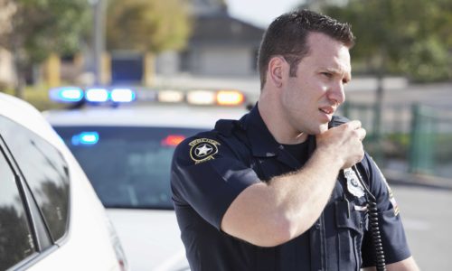 Male police officer (20s) standing outdoors, reaching for radio.  Police car in background.